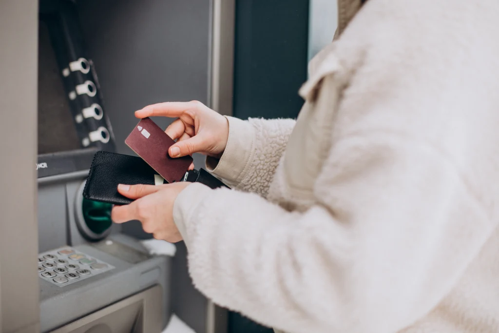 A person in a beige coat uses an ATM, holding a black wallet and a red bank card. The screen and keypad of the ATM are partially visible.