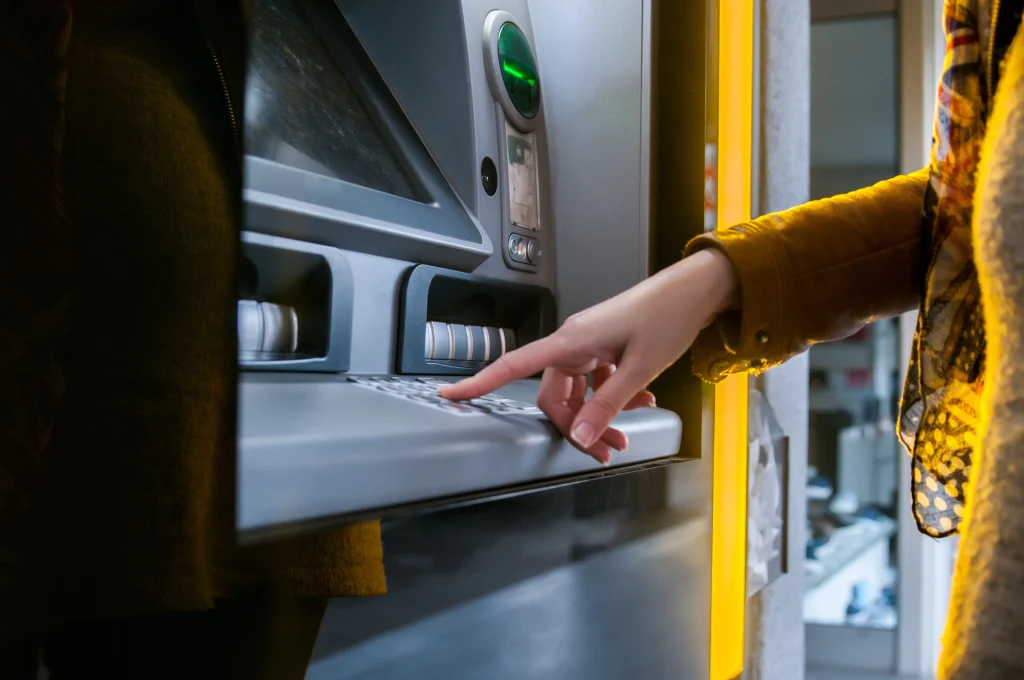A person uses an ATM, pressing buttons on a keypad. The machine glows with yellow light, creating a focused and modern feel.